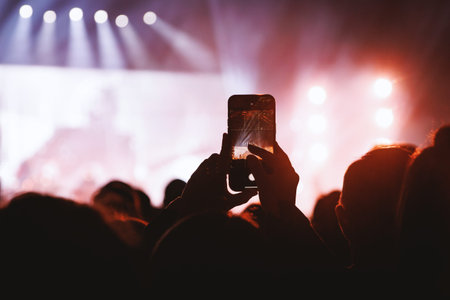 A close-up view of a person's hands holding a smartphone to record a live performance at a music festival under red stage lights.の写真素材