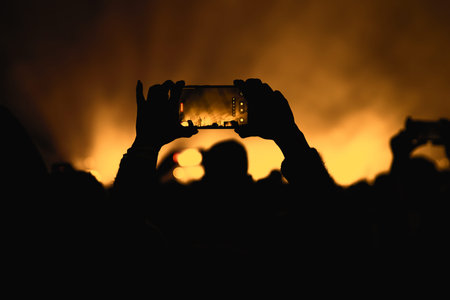 A silhouetted person holding a smartphone to capture a photo of a stage illuminated by bright yellow and golden lights.の写真素材