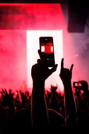 A vertical POV shot of hands holding smartphones to record a live music festival under intense red stage lighting.の写真素材
