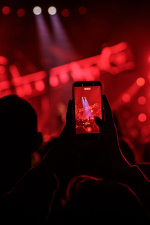A smartphone screen displays a guitarist on stage, framed by intense red lighting and beams. The foreground shows the dark silhouette of the audience.の写真素材