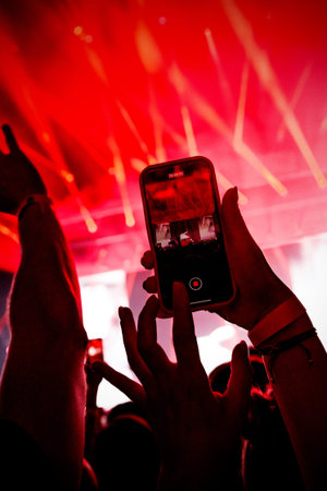 A close-up shot of a mobile phone screen recording a concert performance, illuminated by intense red lighting. The image captures the modern way of documenting live events.の写真素材