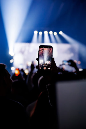A single hand raises a mobile phone to record a live performance. Striking blue spotlights beam down...