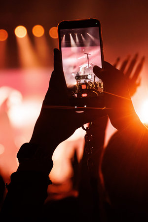 Hands hold a mobile phone horizontally, capturing a video of a singer performing on stage. Bright...