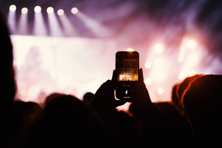 A vertical smartphone is held up by a fan to capture a photo of a brightly lit stage. The background...