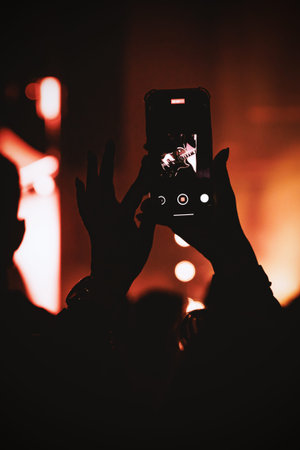 Dramatic silhouette of hands holding a phone in vertical orientation. The screen shows a musician under deep red and orange stage lighting.の写真素材