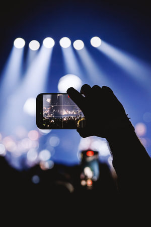 A vertical shot featuring the silhouette of a hand holding a mobile phone to record a live show....