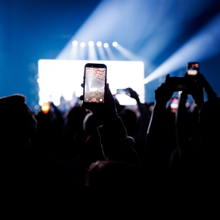 Fans at a live event are silhouetted against an intensely bright white stage screen. One fan in the...