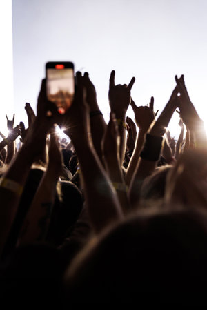 Immersive experience of a live music festival showing silhouettes of a cheering crowd with raised hands and rock signs. Bright stage backlighting creates a dynamic fan perspective.の写真素材