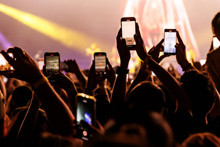A wide view of a concert crowd with numerous glowing smartphone screens held up to record the stage....