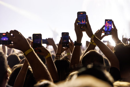 Multiple hands holding up smartphones to film a musician on stage at a large music festival with...