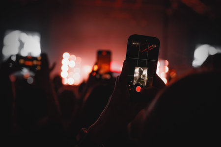 A close-up view of a person's hand holding a mobile phone to record a show. The stage in the...