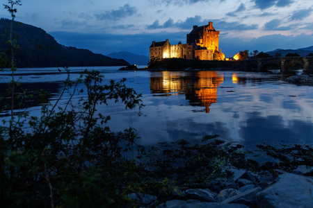 The historic Eilean Donan Castle is illuminated at night and reflected in the dark water of the surrounding loch.の写真素材