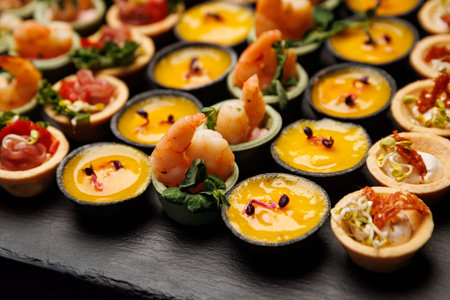 A detail shot of individual party snacks including prawns with greens and yellow savory mousse cups. The appetizers are neatly aligned on a dark stone surface.の写真素材