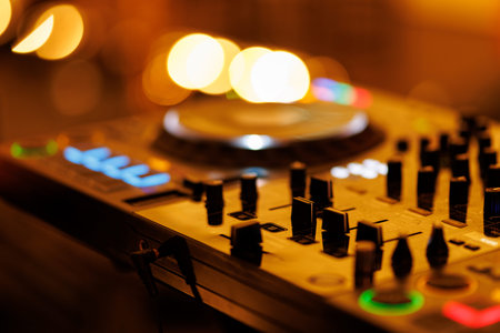 A close-up, low-angle shot of a digital DJ mixing console with various knobs and faders. The background is filled with warm, out-of-focus golden lights.の写真素材