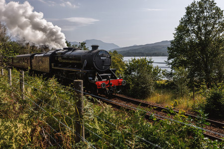 A black steam locomotive emitting white smoke travels along a track near a lake.