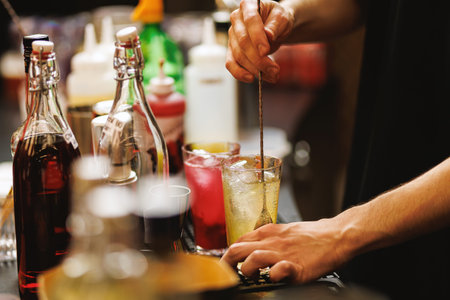 Close-up view of a bartender's hands stirring a drink in a glass surrounded by bottles and bar...