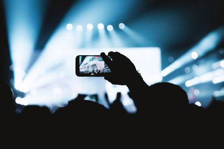 Silhouette of a fan filming a live music performance on a mobile device under dramatic blue light...