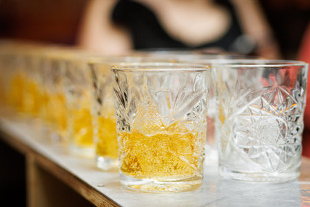 A line of decorative glass tumblers filled with an amber alcoholic beverage stands on a light...