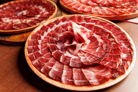 Circular wooden platters are covered in thinly sliced cured ham arranged in a decorative pattern for...