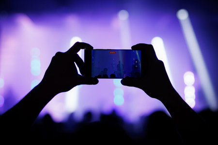 A silhouette of hands holding a mobile phone to film a musical performance on stage under bright...