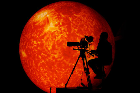 A professional cameraman sits with a tripod-mounted camera silhouetted against a glowing orange...