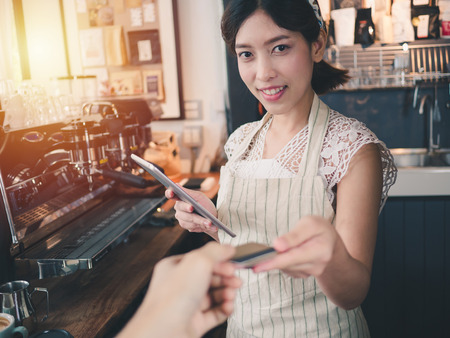 Young asian woman barista smiling standing behind the counter at coffee shop, Using tablet and customer payment by credit card, Cafe restaurant service concept,Owner small business concept.の写真素材