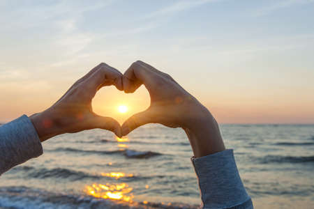 Hands and fingers in heart shape framing setting sun at sunset over ocean Hands and fingers in heart shape framing setting sun at sunset over ocean