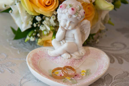 Two beautiful gold wedding rings lie on a platter in a rose shape with the angel sculpture near the bride's bouquet of orange roses and white flowers.の写真素材
