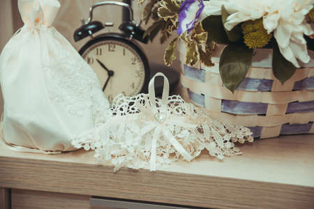 Beautiful lace wedding garter and white handbag of the bride are us the table near the alarm clock.の写真素材