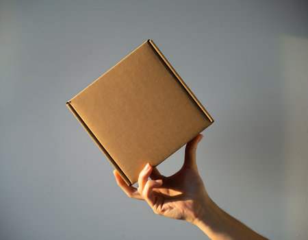 A hand holds a brown cardboard box in front of a gray background, showcasing a simple and minimalist aesthetic.の素材