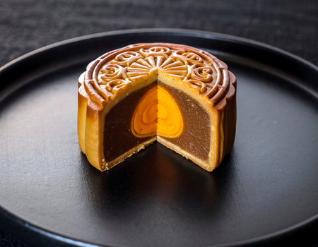 A close-up shot of a cut mooncake, revealing its rich filling and egg yolk center, presented on a sleek black plate.の素材