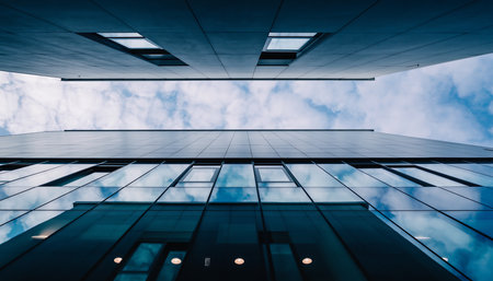 A low-angle perspective of a modern building with glass windows reflecting the sky, showcasing contemporary architectural design and urban aesthetics.の素材
