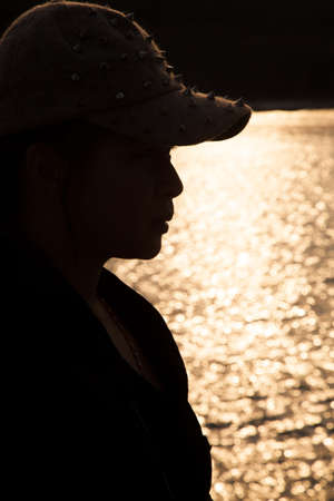 Backlit silhouette of a woman wearing a hat. Sop freckled sunlight reflecting off the surface of the sea in the evening.の写真素材
