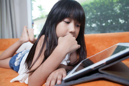 Girl playing with tablet. Lying on the orange sofa at home.の写真素材