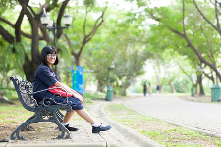 Girl wearing glasses sitting on the bench. Relax under large tree And along corridor within the park.の写真素材