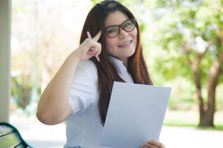 Student wearing glasses, holding a document. Sitting on a bench in the park.の写真素材