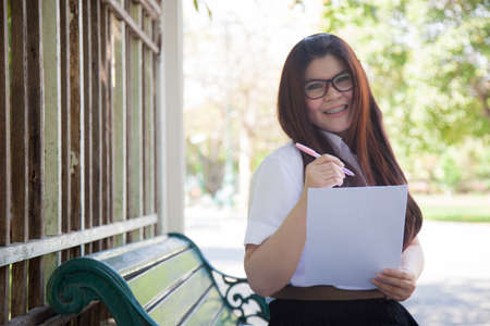 Student wearing glasses, holding a document. Sitting on a bench in the park.の写真素材