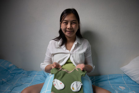 A woman sitting on bed holds green baby outfit with booties, smiling happily. room has soft, cozy...