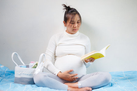 A pregnant woman reading book while sitting on bed, looking thoughtful and serene. scene conveys sense of calm and anticipationの写真素材