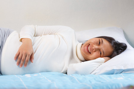 A smiling woman lies on bed, showcasing her pregnancy in cozy setting. soft colors and relaxed pose create warm, inviting atmosphereの写真素材