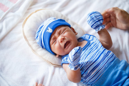 A Crying baby in blue outfit with hat and mittens on soft beddingの写真素材