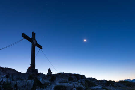mountain cross on peak with moon at blue sky sunrise