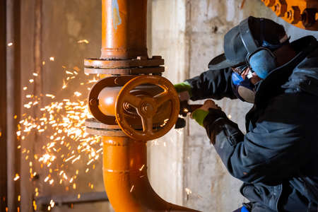 worker man with breathing mask and ear protection flexes with an angle grinder on orange steel pipes with flying sparksの写真素材