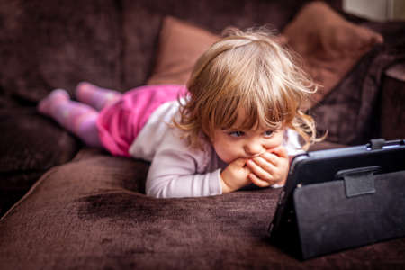 Little girl laying on a sofa and watching cartoon on a small tabletの写真素材