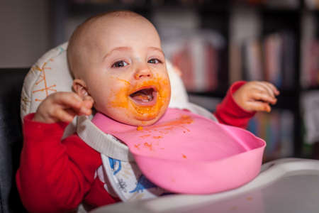 Little boy sitting in a high chair at home and getting his face dirty while eating his lunchの写真素材