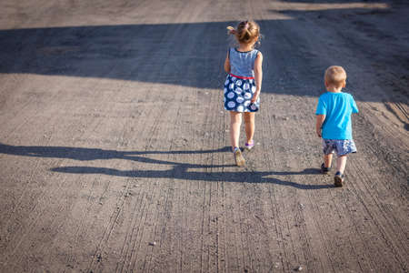 Little boy and his older sister walking on a gravel, dusty and corrugated village road in the...