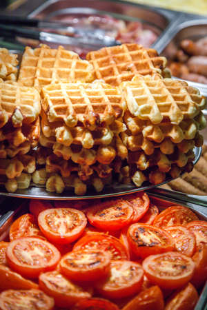 Hot counter containing warm breakfast items such as scrambled or fried tomatoes, waffles, bacon, sausages and others in a self service restaurant in a hotelの写真素材