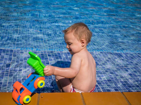 Cute two years old boy having fun and playing with plastic toy car in water by the outdoor swimming poolの写真素材