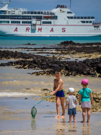 Little boy and girl standing on the beach and watching older boy with a net catching little crabs, passenger ferry Armas in the background.  Picture taken11 April 2016 in Corralejo, Fuerteventura, Spainのeditorial素材