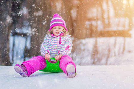 Portrait of a little girl sitting on a green plastic sled in the park in winterの写真素材